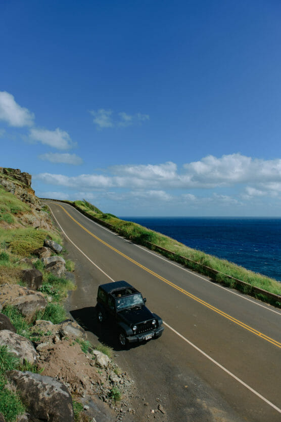 A black SUV is parked on the side of a winding coastal road with green hills and rocks on one side and the deep blue ocean under a bright blue sky with scattered clouds on the other. by Justin Salem Meyer