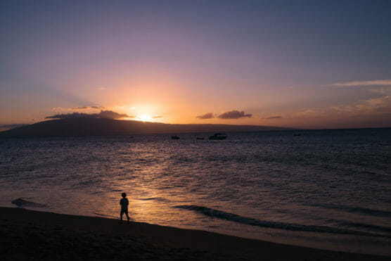 A child walks along the beach at sunset, with the sun setting behind a distant mountain and boats silhouetted on the calm ocean. The sky is filled with warm orange and purple hues. by Justin Salem Meyer