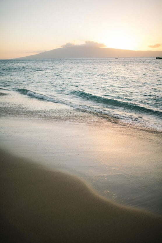 Gentle waves wash onto a sandy beach at sunset, with soft golden light reflecting on the water. In the distance, a mountain is silhouetted against the sky as the sun sets behind it. by Justin Salem Meyer