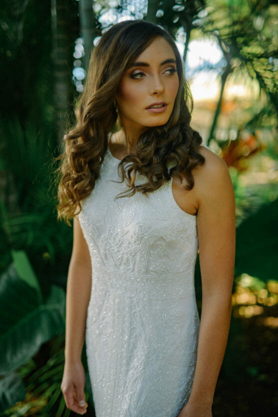 A woman wearing a white, intricately patterned sleeveless dress stands outdoors among lush green foliage, with long, wavy brown hair and a thoughtful expression, looking slightly to the side. by Justin Salem Meyer