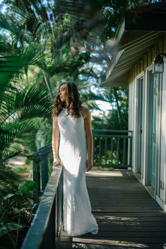 A woman in a white sleeveless dress stands on a wooden porch surrounded by lush green plants, gazing thoughtfully into the distance with sunlight filtering through the leaves. by Justin Salem Meyer