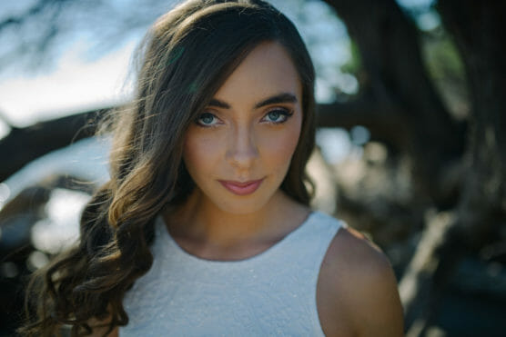 A young woman with long brown hair and blue eyes, wearing a sleeveless white top, looks directly at the camera outdoors with blurred trees in the background. by Justin Salem Meyer