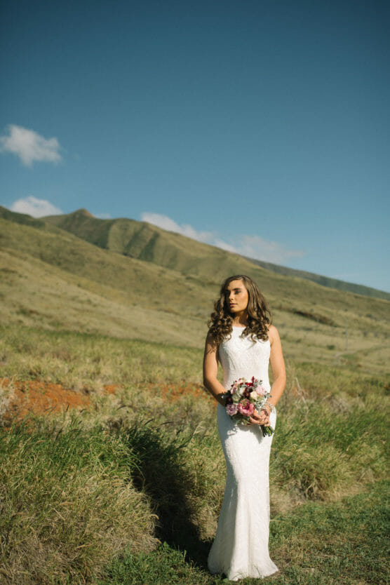 A bride in a white dress holding a bouquet of flowers stands in a grassy field with green hills and a blue sky in the background. by Justin Salem Meyer