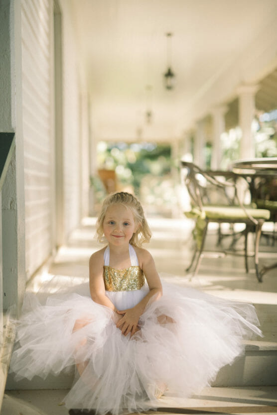 A young girl in a white tutu dress with a gold sequined top sits on porch steps, smiling softly. Sunlight filters through the porch, illuminating her and the airy fabric of her dress. by Justin Salem Meyer