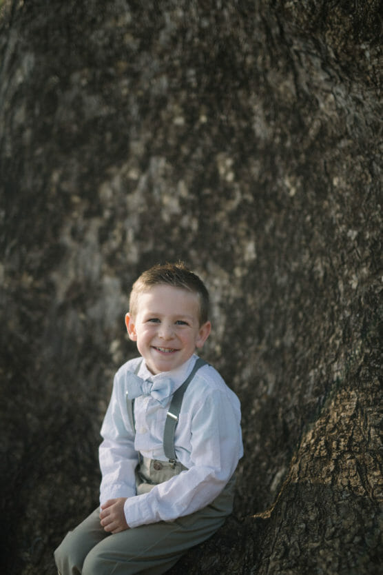 A young boy in a white shirt, bow tie, and suspenders sits and smiles in front of a large tree trunk. by Justin Salem Meyer