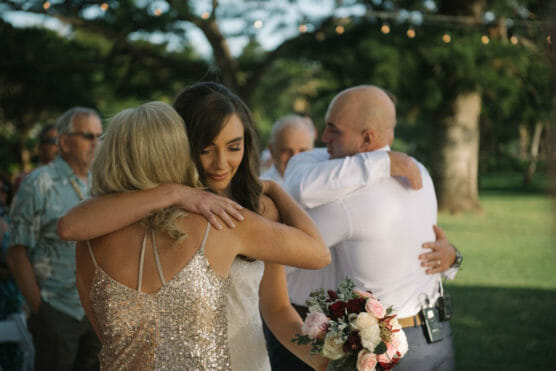 A bride in a white dress hugs a woman in a sparkly dress, while two men embrace in the background at an outdoor wedding. The bride holds a bouquet of pink and white flowers. Trees and guests are visible behind them. by Justin Salem Meyer