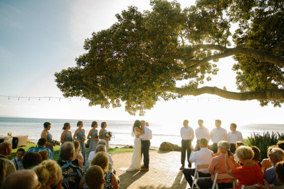 A bride and groom kiss under a large tree during an outdoor wedding ceremony by the ocean, with bridesmaids, groomsmen, and seated guests looking on. The sun shines brightly in the background. by Justin Salem Meyer