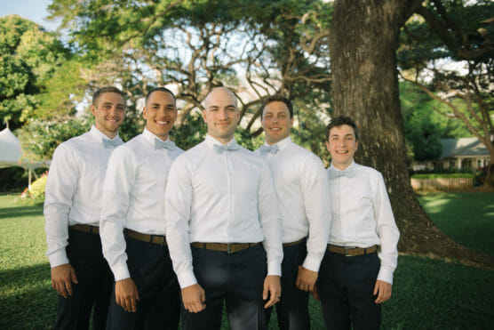 Five men wearing white shirts and bow ties stand together outdoors on grass, smiling in front of a large tree with sunlight filtering through the branches. by Justin Salem Meyer