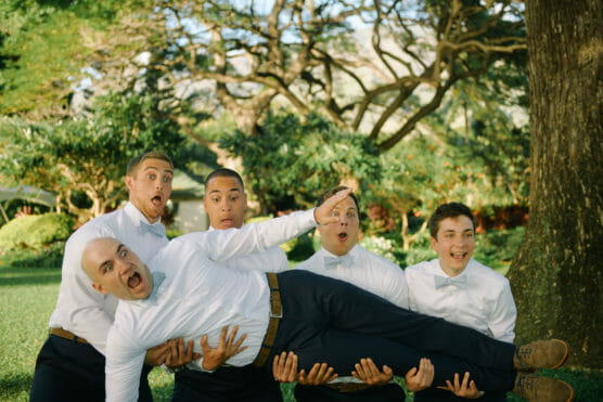 Five men in white shirts and bow ties stand outdoors. Four of them are holding up the fifth man horizontally. All have surprised or playful expressions. Lush greenery and trees fill the background. by Justin Salem Meyer