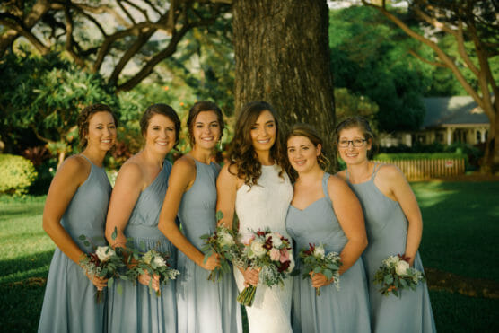 Six women posing outdoors in front of a large tree, with five wearing light blue dresses holding bouquets and one in a white bridal gown, all smiling at the camera. Lush greenery and a house are in the background. by Justin Salem Meyer