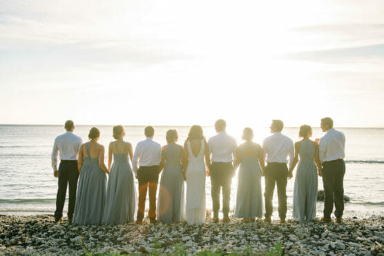 A group of people dressed in formal attire, with women in light gray dresses and men in white shirts and dark pants, stand side by side facing the ocean at sunset on a rocky beach. by Justin Salem Meyer