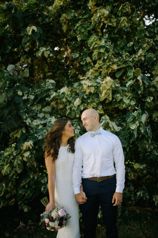 A bride in a white dress holding a bouquet stands beside a groom in a white shirt and bow tie. They smile at each other, holding hands, in front of lush green foliage. by Justin Salem Meyer