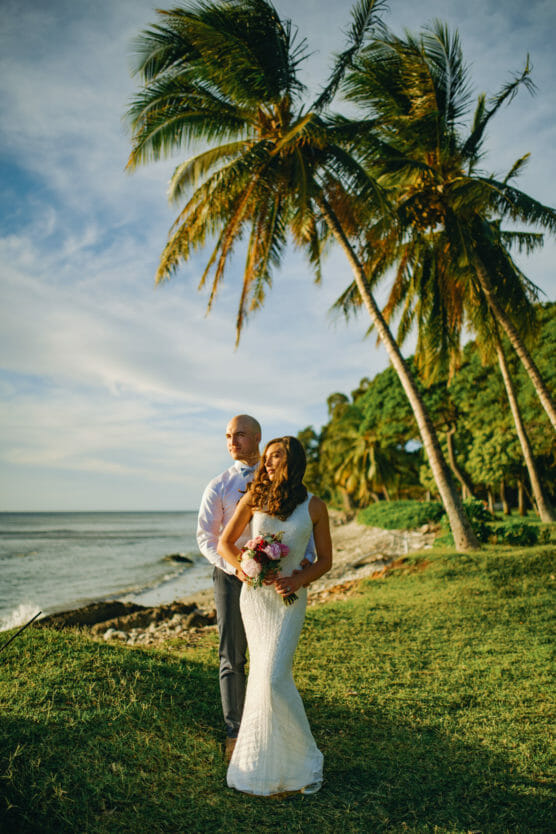 A bride in a white dress holding a bouquet stands in front of a groom in a white shirt and tie, with palm trees and the ocean in the background under a sunny sky. by Justin Salem Meyer