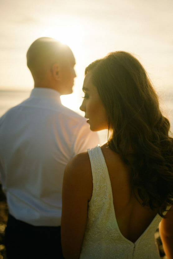 A woman in a white dress stands in the foreground, looking to the side, while a man in a white shirt stands slightly ahead of her, both bathed in warm, golden sunlight. by Justin Salem Meyer
