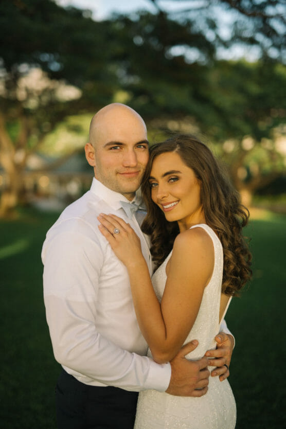 A couple in formal attire embraces outdoors, smiling at the camera. The woman wears a white dress and shows an engagement ring; the man is in a white shirt. They stand on green grass with trees in the background. by Justin Salem Meyer