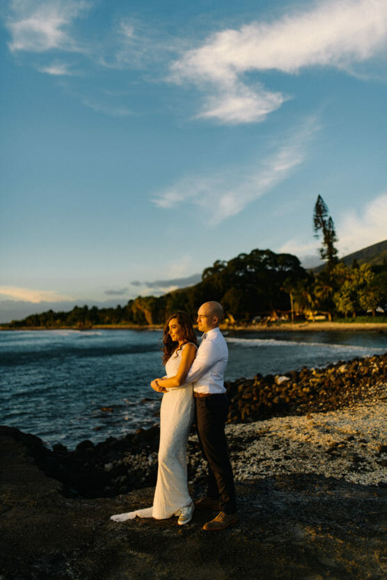 A couple in wedding attire stands on a rocky beach at sunset, with the groom embracing the bride from behind. The sky is partly cloudy and the coastline, trees, and mountains are visible in the background. by Justin Salem Meyer