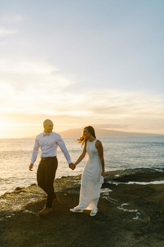 A couple holds hands and walks along a rocky shoreline at sunset. The woman wears a white dress and the man is in a white shirt and dark pants. The ocean and a distant island are visible under a colorful sky. by Justin Salem Meyer