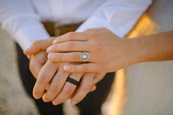 Close-up of two hands gently holding each other, one hand wearing a diamond engagement ring and the other a black wedding band, both dressed in light-colored clothing. by Justin Salem Meyer