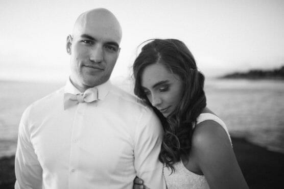 A bride rests her head on the groom’s shoulder as they stand together by the water. The groom wears a white shirt and bow tie, while the bride wears a white dress. The image is in black and white. by Justin Salem Meyer