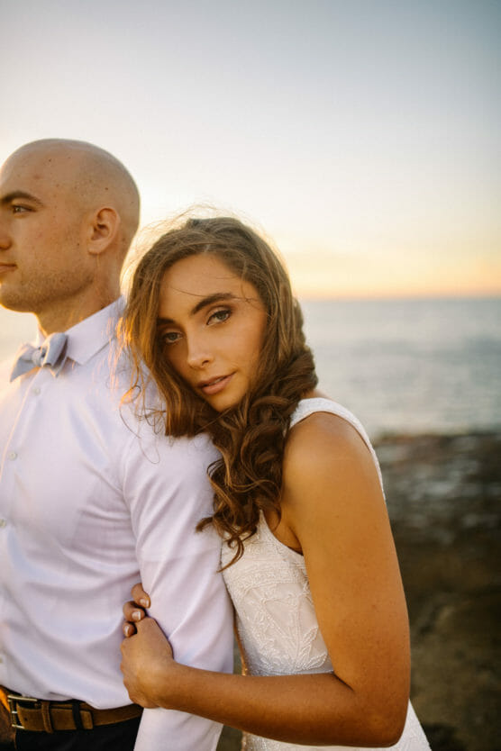 A woman in a white dress leans her head on the arm of a man in a white shirt and bowtie, standing close together by the ocean at sunset. She looks at the camera while he looks away. by Justin Salem Meyer