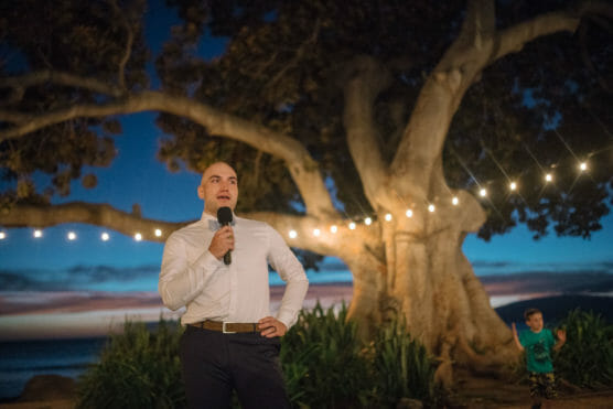 A man holding a microphone stands under string lights near a large tree at dusk, with a sunset and ocean in the background. A child in a blue shirt is visible to the right. by Justin Salem Meyer