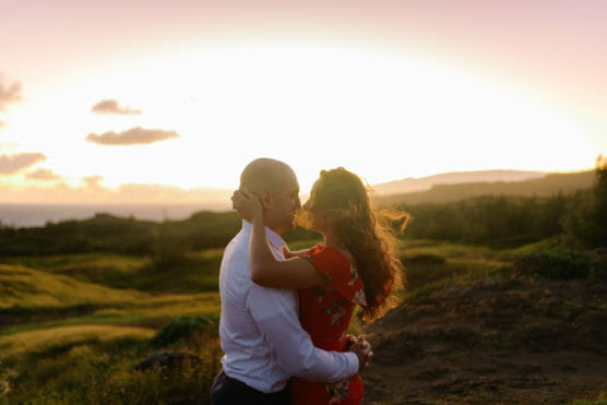 A couple embraces and kisses outdoors at sunset, with soft sunlight illuminating the landscape and hills in the background. The woman wears a red dress, and the man wears a white shirt. by Justin Salem Meyer