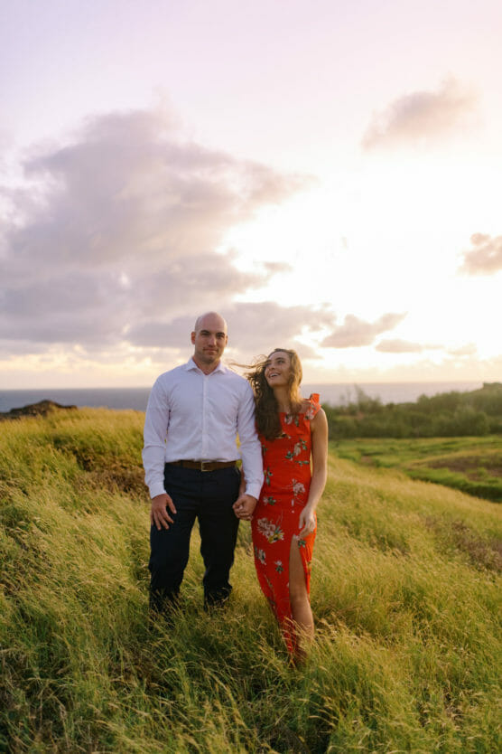 A man in a white shirt and dark pants stands holding hands with a woman in a red floral dress, smiling in a grassy field at sunset with a cloudy sky in the background. by Justin Salem Meyer