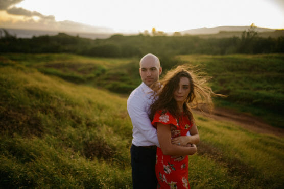 A man and woman stand in a grassy field at sunset. The man in a white shirt stands behind the woman in a red floral dress, with the woman’s hair blowing in the wind. The landscape is lush and softly lit. by Justin Salem Meyer