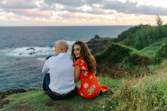 A man and woman sit on grassy cliffs overlooking the ocean at sunset. The woman, in a red floral dress, looks back and smiles at the camera while the man, in a white shirt, gazes at the sea. by Justin Salem Meyer