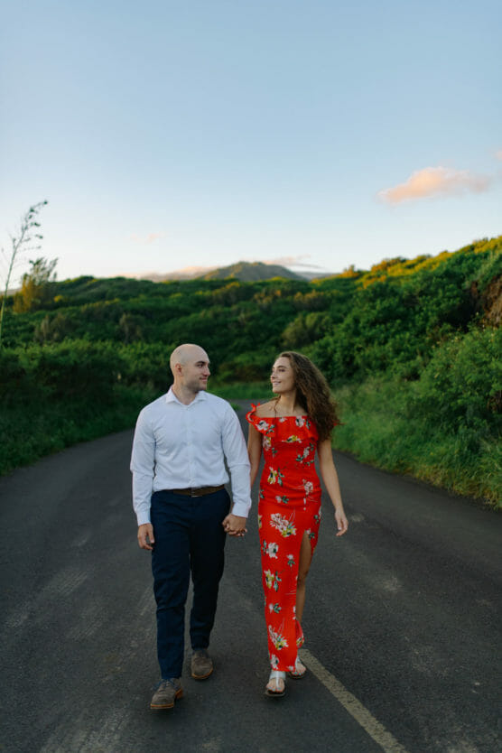 A couple walks hand in hand down a paved road surrounded by lush green hills. The man wears a white shirt and navy pants; the woman wears a red floral dress. They look at each other and smile under a clear sky. by Justin Salem Meyer