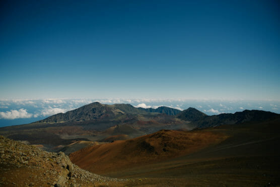 View of a volcanic landscape with red and brown rocky terrain in the foreground and a mountain ridge in the distance under a clear blue sky above a layer of clouds. by Justin Salem Meyer
