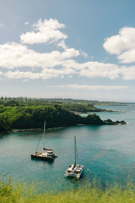 Two sailboats float on calm blue water near a lush, green coastline under a partly cloudy sky, with rolling hills and trees in the background. by Justin Salem Meyer