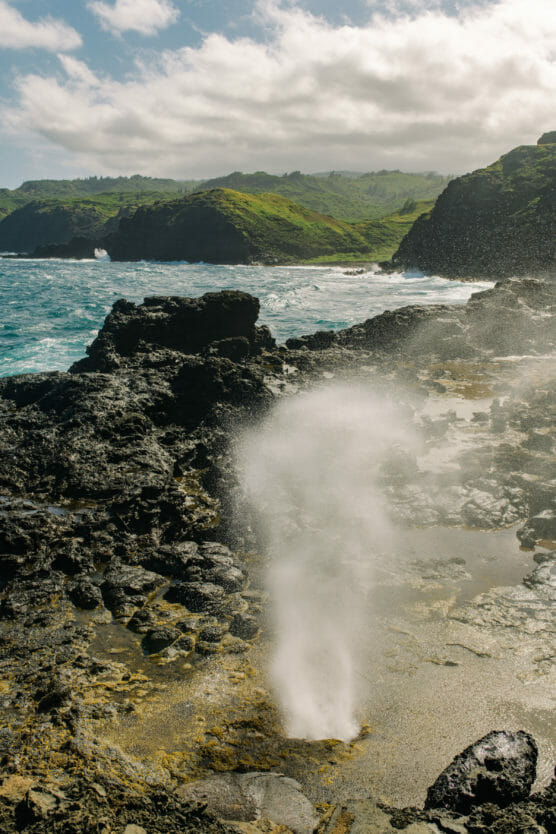A coastal scene with rugged black rocks and ocean waves. A spout of water sprays upward from a blowhole in the foreground. In the background, green hills rise under a partly cloudy sky. by Justin Salem Meyer