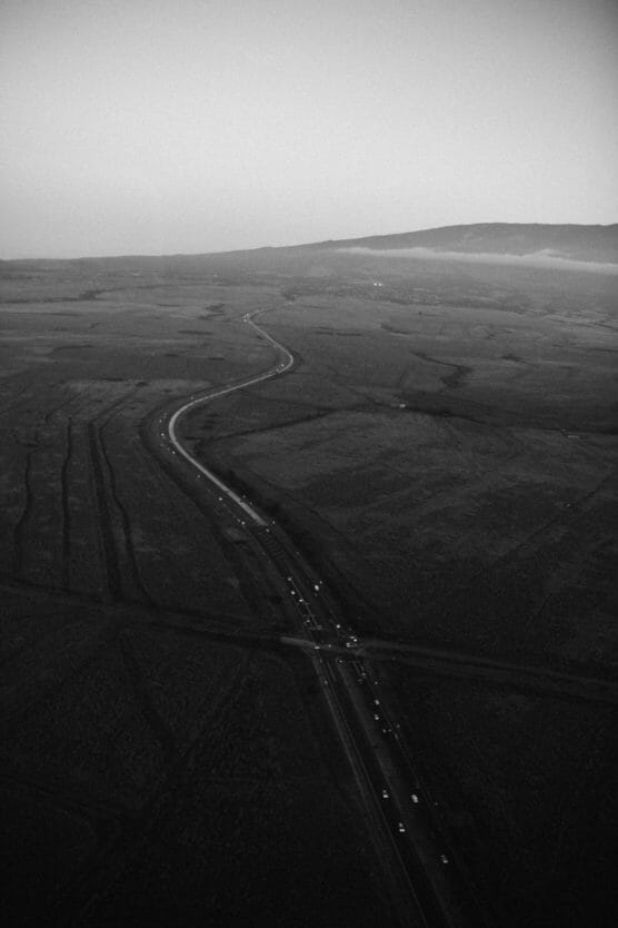 A winding road cuts through a vast, open landscape under a hazy sky, with distant hills in the background and small vehicles visible along the road. The scene is in black and white. by Justin Salem Meyer