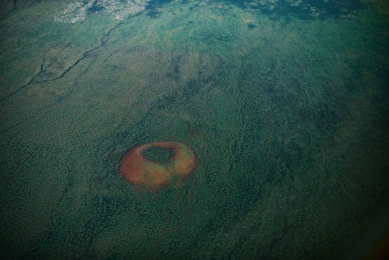 Aerial view of a large, circular volcanic crater with reddish edges surrounded by green landscape, with scattered vegetation and a lighter area near the top of the image. by Justin Salem Meyer