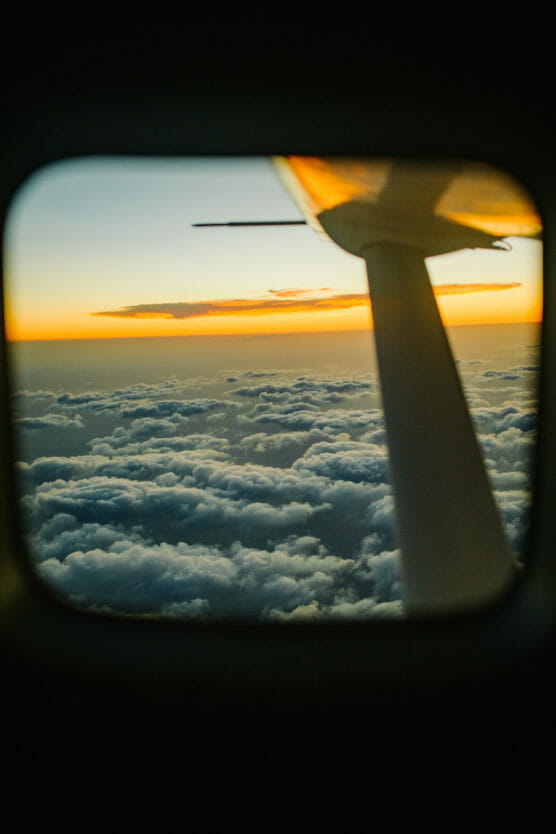 View from an airplane window showing a wing, fluffy clouds below, and a vibrant orange and yellow sunset sky above the horizon. by Justin Salem Meyer