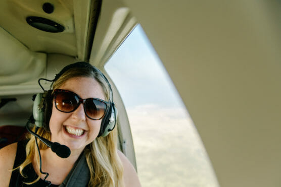A woman wearing sunglasses and a headset smiles widely while sitting inside a small aircraft, with sunlight streaming through the window beside her. by Justin Salem Meyer