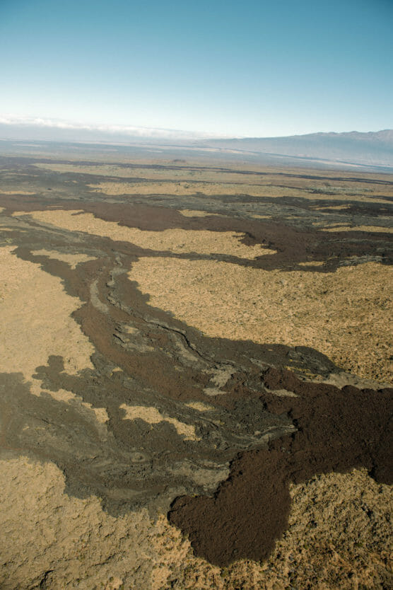 Aerial view of a dry, flat landscape with dark, winding lava flows cutting through light brown desert terrain under a clear blue sky. Mountains are visible in the distant background. by Justin Salem Meyer