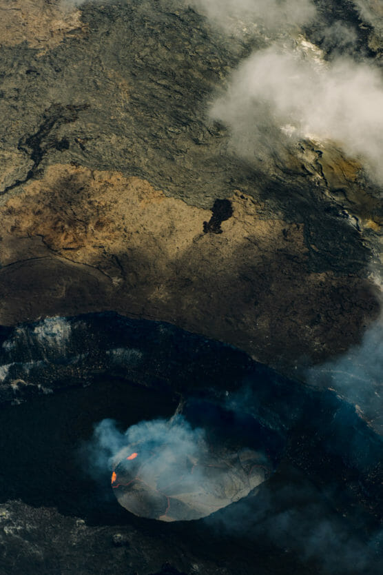Aerial view of an active volcanic crater with visible flowing lava, surrounded by dark, cracked earth and rising white steam or smoke. by Justin Salem Meyer