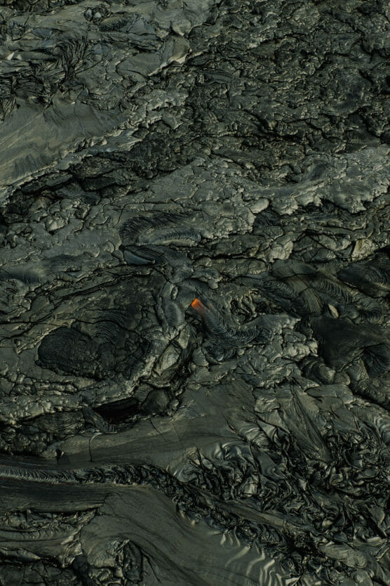 A close-up view of dark, cooled lava with rough, textured surfaces and a small, glowing patch of orange molten lava visible near the center. by Justin Salem Meyer