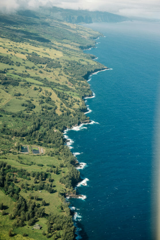 Aerial view of a lush green coastline with rocky cliffs and waves crashing along the shore, meeting deep blue ocean water under a partly cloudy sky. Farmland and forest areas are visible inland. by Justin Salem Meyer