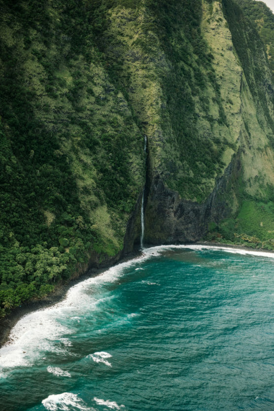 A tall waterfall cascades down a steep, green cliff into the ocean, with waves crashing along a rocky shoreline below. by Justin Salem Meyer