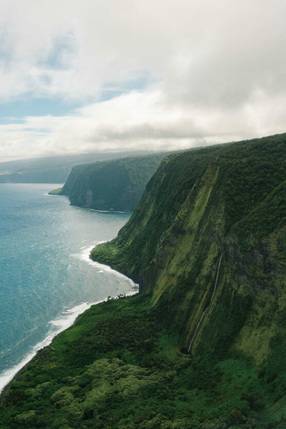 Aerial view of lush green cliffs meeting the blue ocean, with a narrow waterfall cascading down one cliff and misty clouds overhead. by Justin Salem Meyer