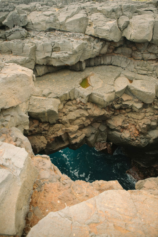 Large, rugged volcanic rock formations with a natural opening reveal bright blue ocean water below; some small puddles of water sit atop the rocks. by Justin Salem Meyer
