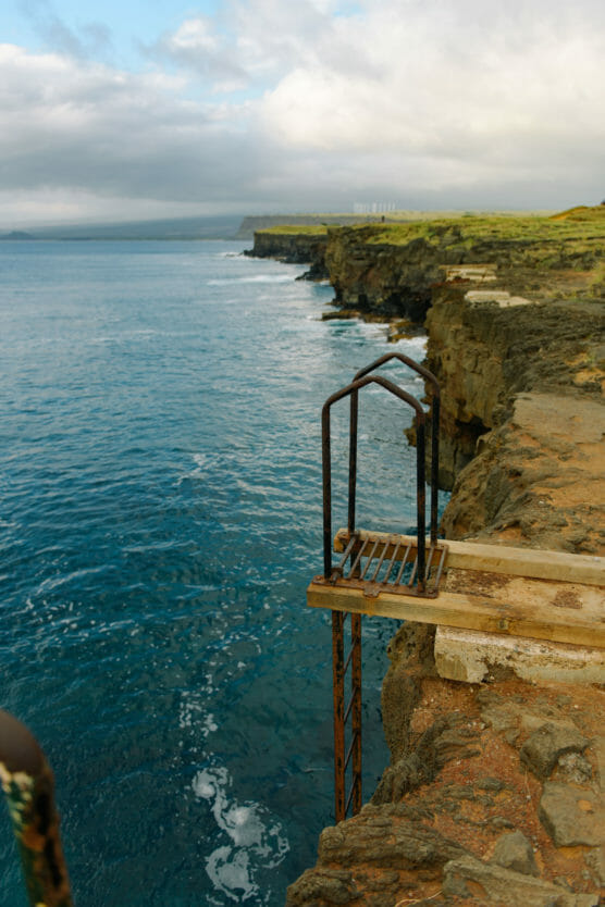 A rusty metal ladder attached to a rocky cliff edge leads down to turquoise ocean water, with dramatic cliffs and grassy land stretching into the distance under a cloudy sky. by Justin Salem Meyer