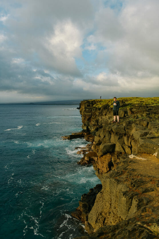 A person stands on the edge of a rocky cliff overlooking the ocean, with blue water below, waves crashing against the rocks, and a cloudy sky above. Another person is visible in the distance on the cliff. by Justin Salem Meyer