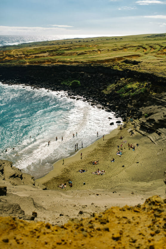 People relax and swim on a sandy beach bordered by turquoise ocean waves and rocky cliffs, with grassy, rolling landscape extending into the distance under a bright sky. by Justin Salem Meyer
