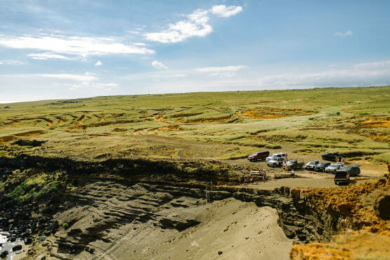 Several vehicles are parked on a grassy, rugged plateau near the edge of a steep rocky cliff, with expansive open fields stretching into the distance under a partly cloudy sky. by Justin Salem Meyer