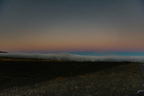A scenic view of a dark landscape at dusk with a low, thick layer of fog rolling over distant hills, under a gradient sky transitioning from blue to pink and purple hues. by Justin Salem Meyer