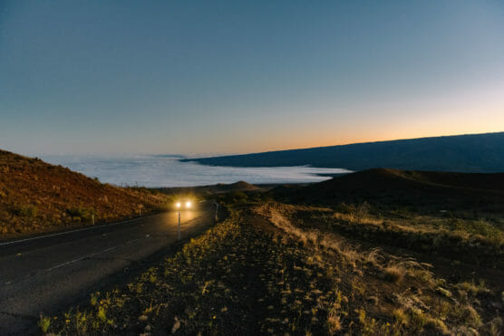 A car with headlights on drives along a winding road at dusk, bordered by grassy hills, with a layer of clouds or fog settled in the distant valley under a clear sky. by Justin Salem Meyer